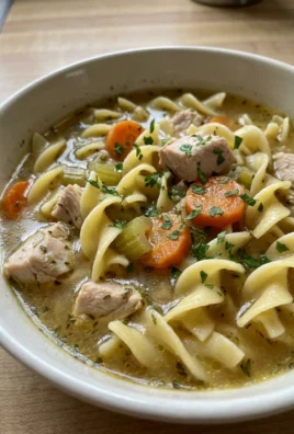 A close-up of a white bowl filled with chicken noodle soup with a spoon.